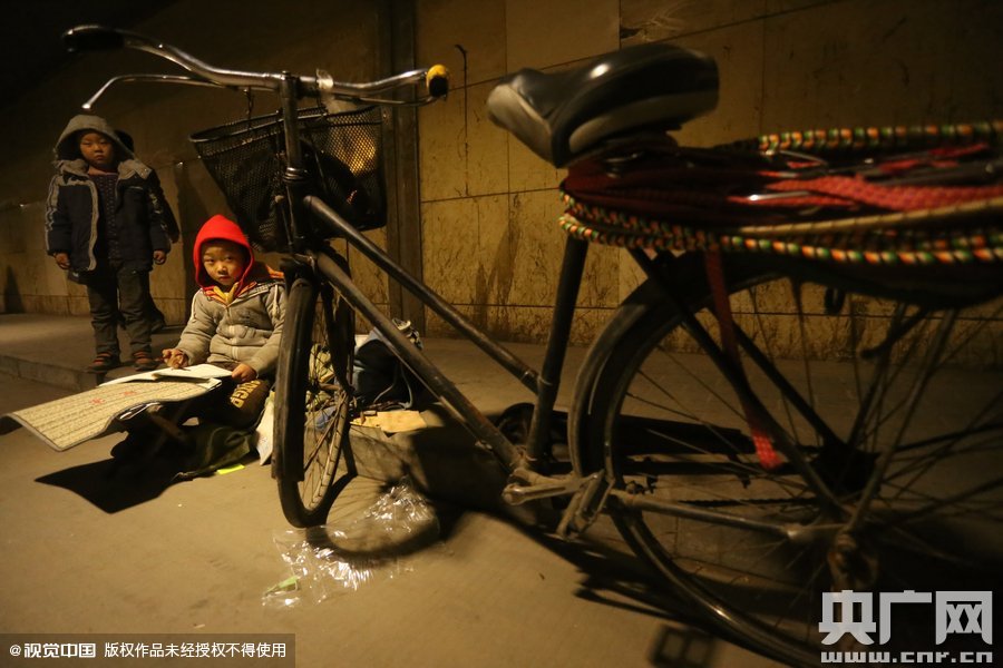 2015年11月8日晚，鄭州，7歲男孩涵洞橋下陪父母練攤，路邊燈下坐小馬扎上寫(xiě)作業(yè)。